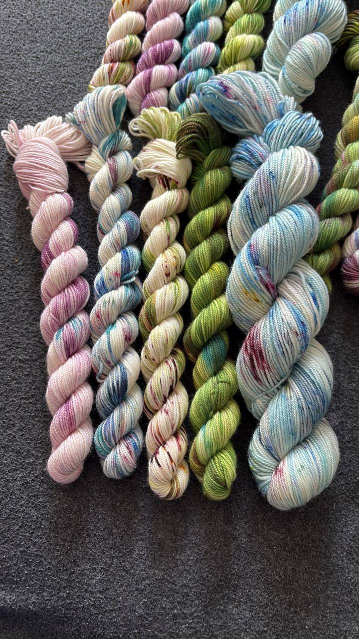 Geverfde strengen garen in roze, blauw, groen en gespikkelde tinten op een donkere ondergrond.