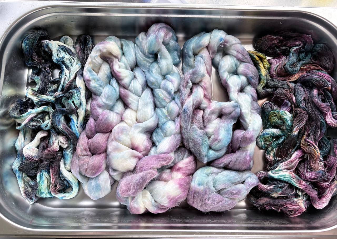 Geverfde vezel en strengen garen in roze, blauw, groen en donkere tinten in een metalen bak.